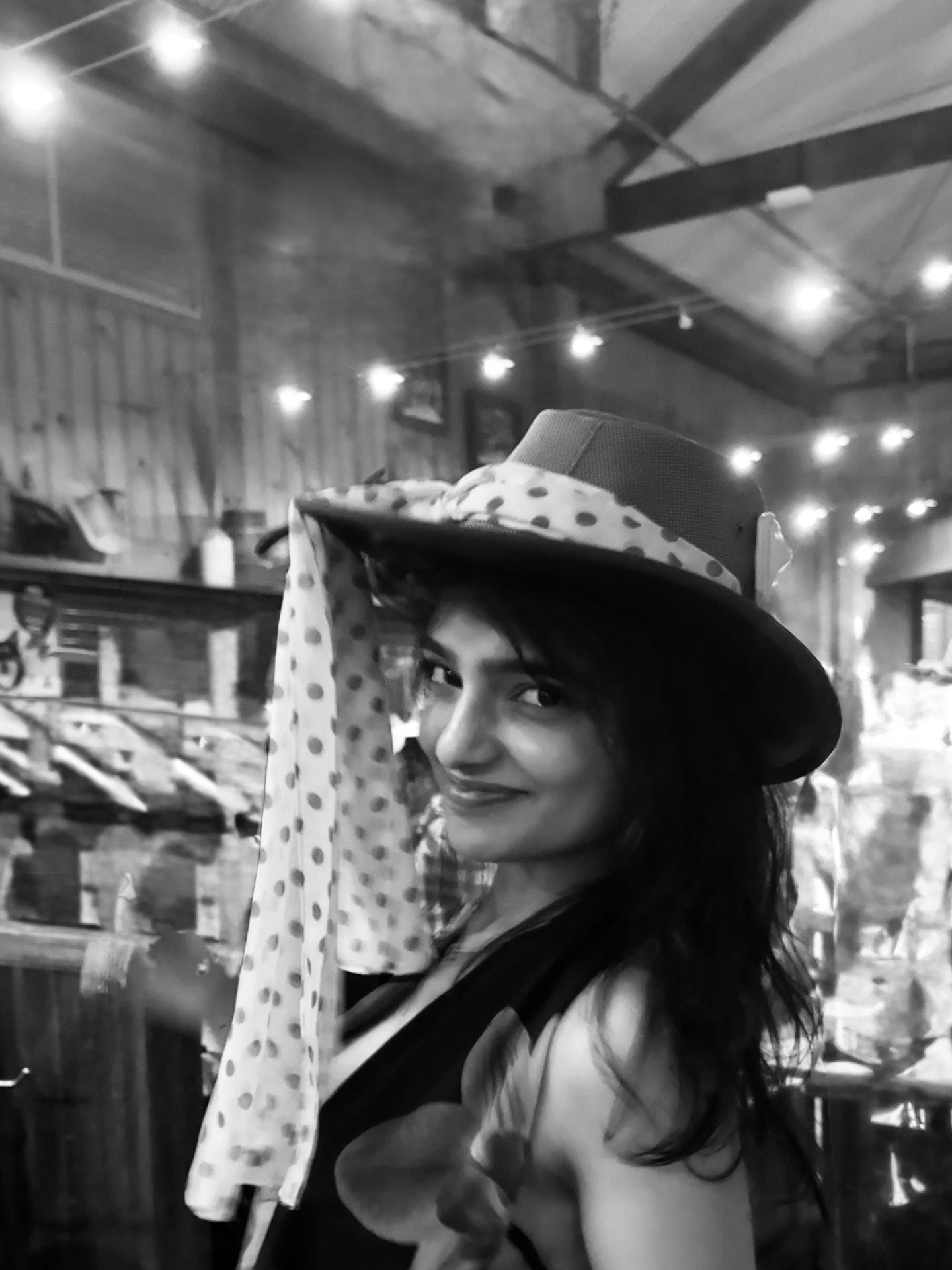 Black and white portrait of Rashika Jain smiling in a wide-brimmed hat under warm string lights